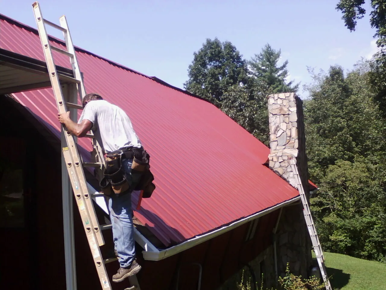 Roofer on a ladder beside a red metal roof and stone chimney — Soffit & Fascia Repair by Roofing Brothers in Centertown